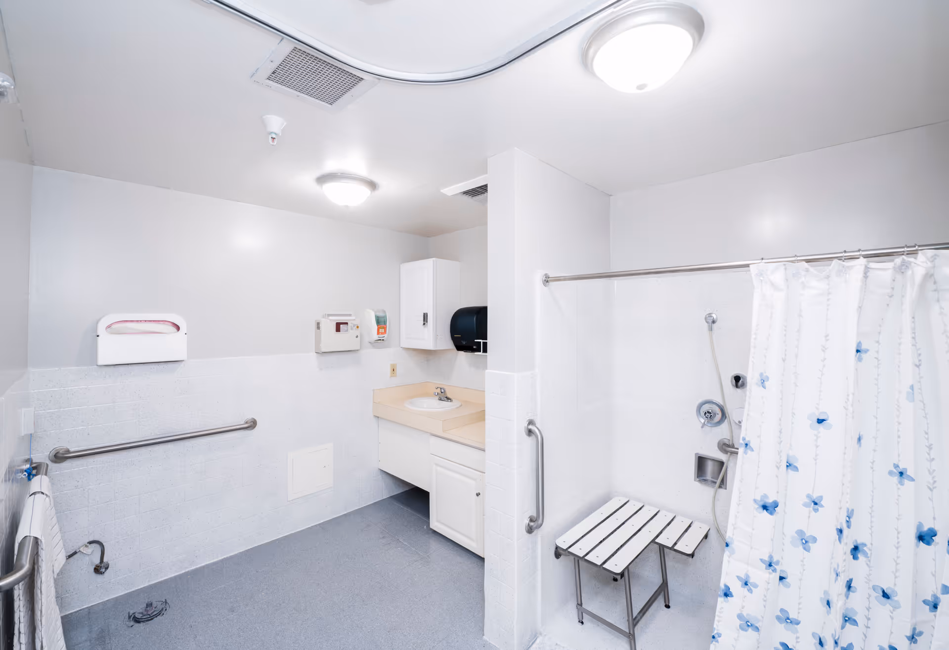 A clean, accessible bathroom with white walls and a gray floor. The room features a sink with a beige countertop, a wall-mounted paper towel dispenser, and a soap dispenser. There are grab bars on the walls and a shower area with a white and blue floral shower curtain and a foldable shower seat.