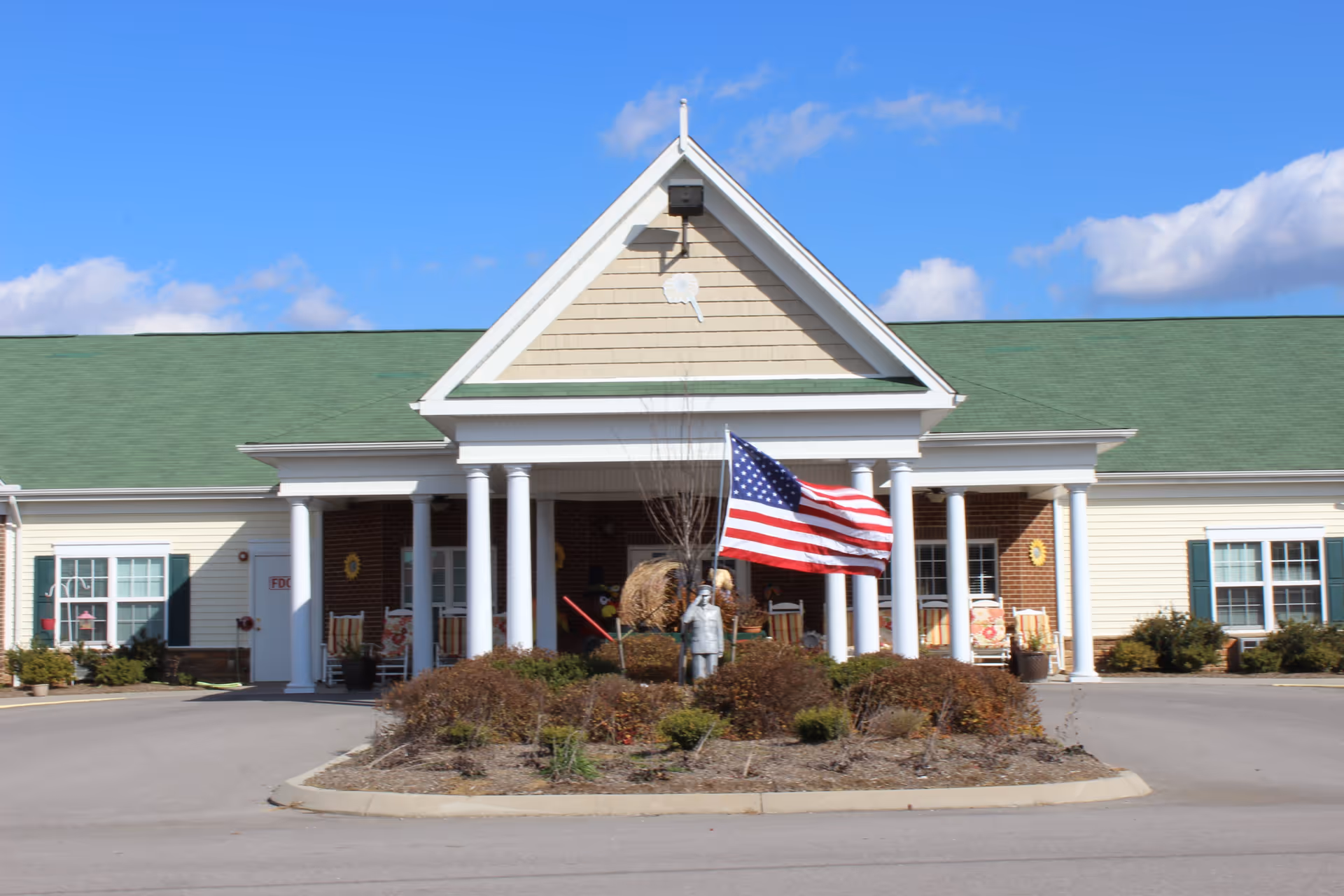 Front exterior view of Meadow View Senior Living facility with a green roof, white columns, and an American flag in the landscaped roundabout in front of the entrance.