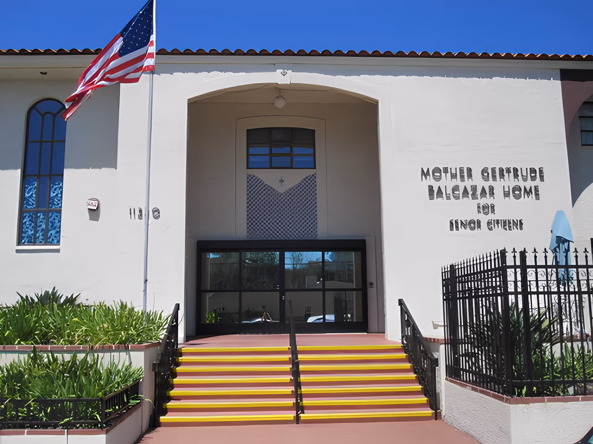 Front entrance of the Mother Gertrude Balcazar Home for senior citizens with yellow-striped steps, an American flag, and signage on the white facade.