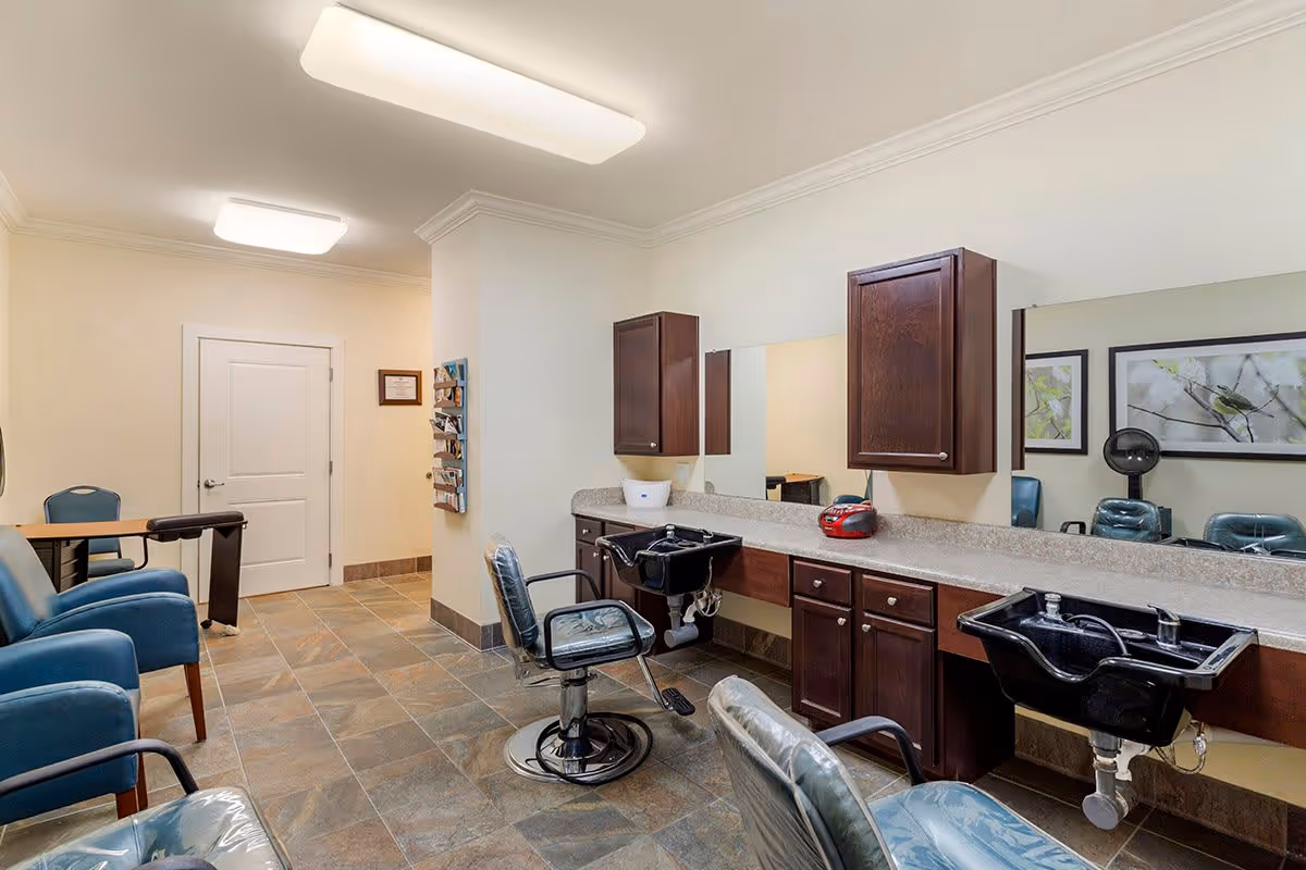 Interior view of a salon area in an assisted living facility with two black hair washing sinks, salon chairs, wooden cabinets, a large mirror, and blue upholstered chairs along the wall. The floor is tiled and the walls are light-colored with framed artwork.