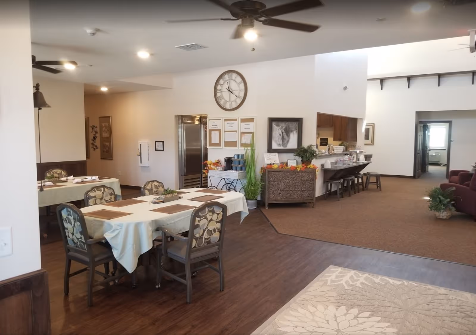 Interior view of a senior living facility dining and common area with tables covered in white tablecloths and chairs around them. There is a large wall clock, a decorative cabinet with plants and fall decorations, a kitchen counter with stools, ceiling fans, and a carpeted lounge area with armchairs in the background.