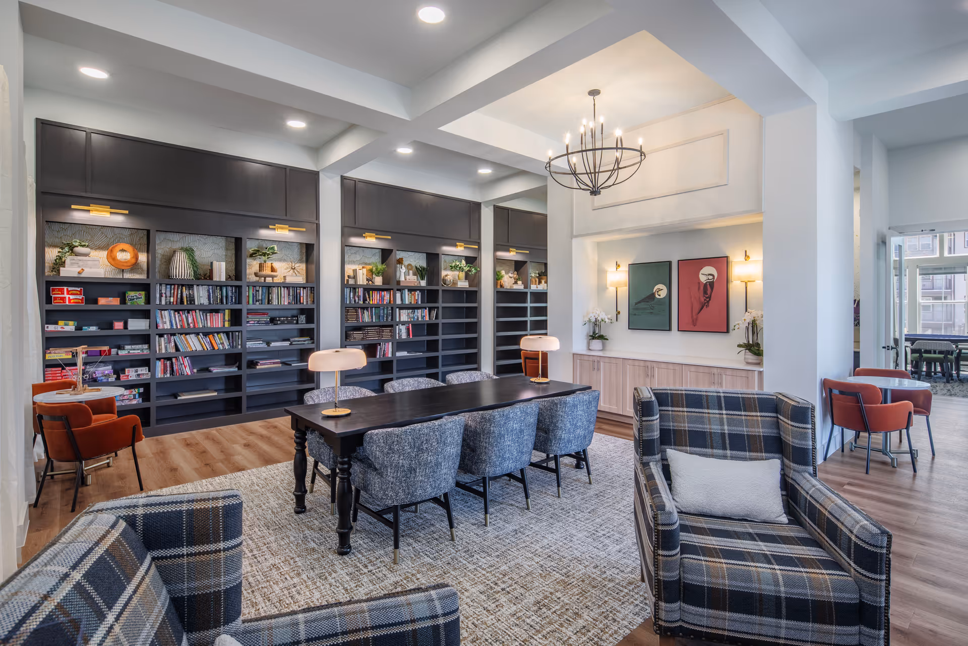 A bright and modern common area featuring a large black table surrounded by six blue upholstered chairs, two plaid armchairs with a small pillow each, and a wall of built-in dark bookshelves filled with books and decorative items. The room has wooden flooring, a beige patterned rug, a chandelier, and two framed artworks on the wall with two wall sconces. In the background, there are additional seating areas with orange chairs and a glass door leading to another room.