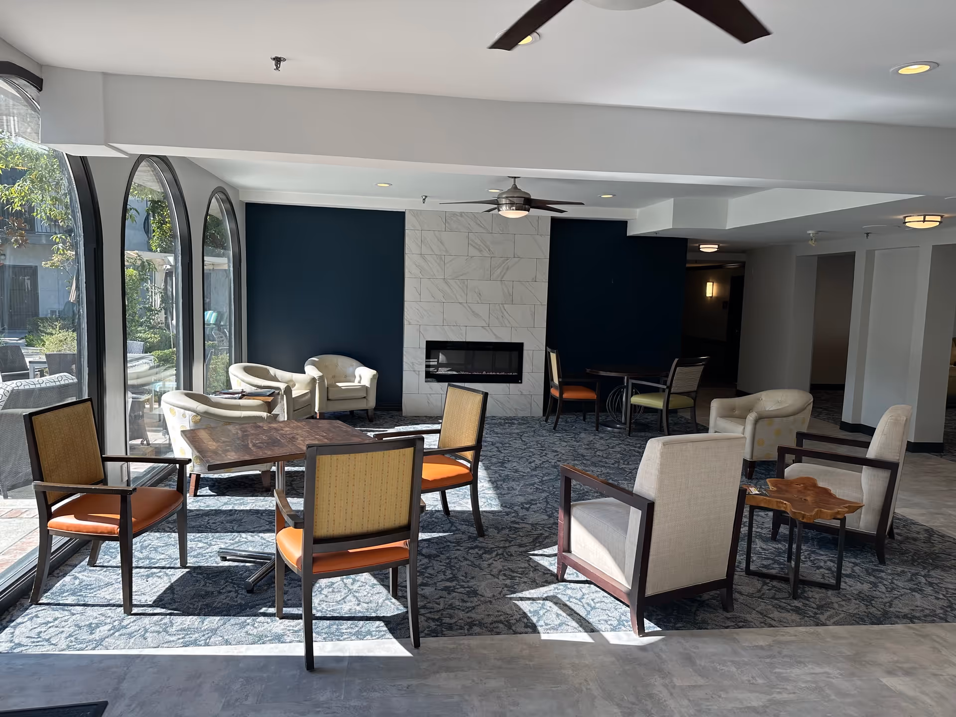A bright and spacious senior living common area with large arched windows letting in natural light. The room features a modern fireplace set in a white tiled wall with dark blue accent walls on either side. There are several seating arrangements including cushioned armchairs with wooden frames, small tables, and a patterned carpet covering the floor. Ceiling fans and recessed lighting are visible on the white ceiling.