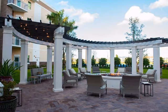 Outdoor seating area with cushioned chairs arranged around a circular fire pit under a pergola structure with string lights. The area is paved with bricks and surrounded by greenery and a multi-story building in the background.