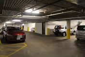 Indoor parking garage with several parked cars, concrete pillars painted with yellow protective padding, and overhead lighting fixtures.