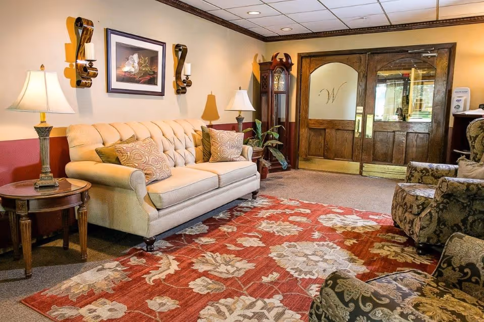 A cozy living room area with a beige tufted sofa adorned with patterned pillows, two floral upholstered armchairs, a wooden side table with a lamp, a grandfather clock, and double wooden doors with frosted glass panels featuring a decorative 'W'. The room has warm lighting and a large red and beige floral patterned rug.