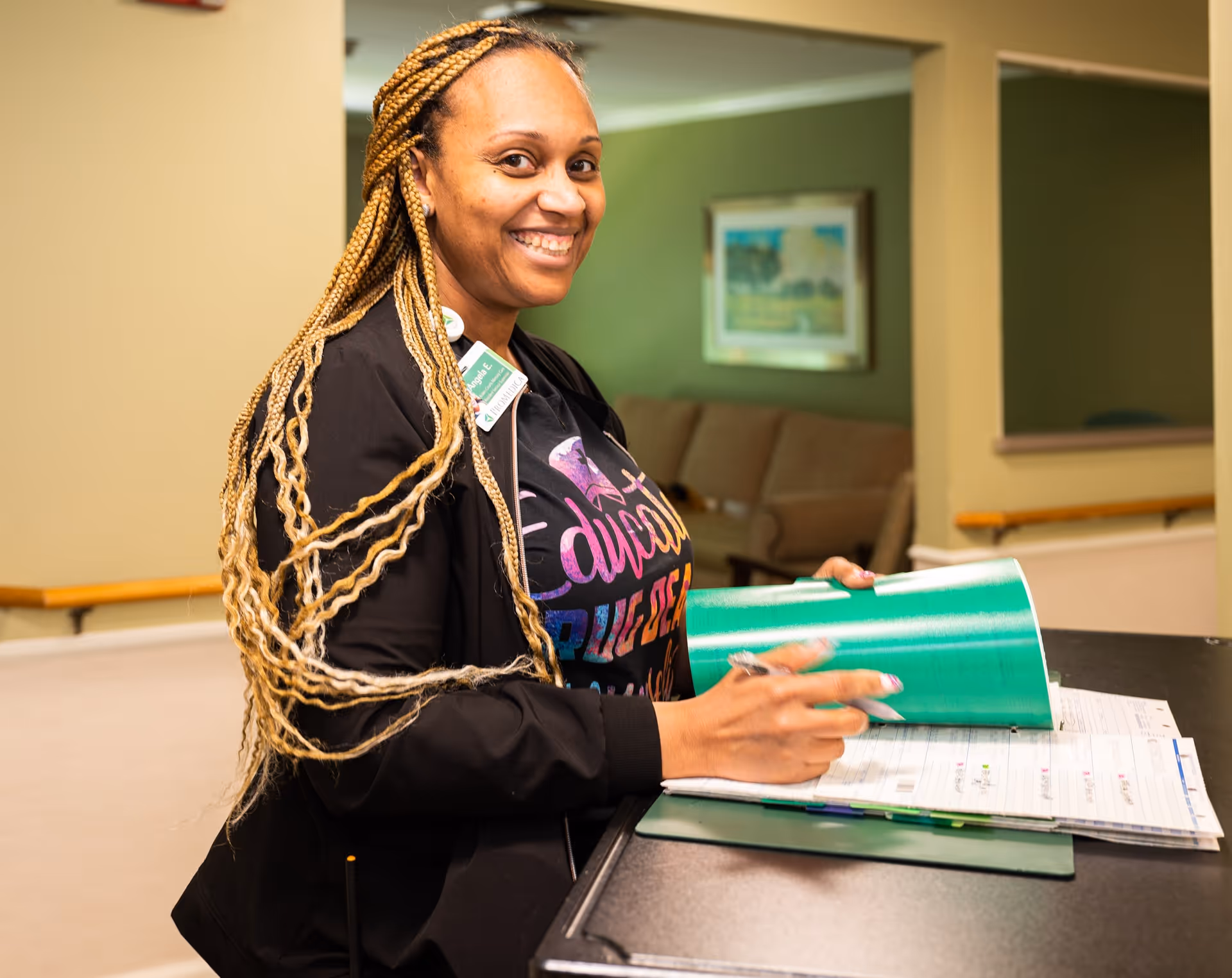 A smiling woman with long braided hair stands at a counter, holding a green folder and pen, with a beige couch and framed artwork visible in the background.