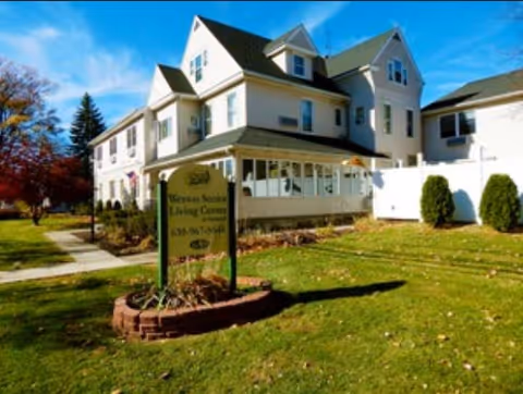 Front exterior of a large white Victorian-style senior living facility with a lawn, walkway, and a sign near the sidewalk.