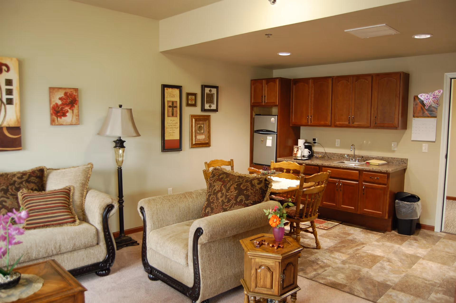 A cozy living area with two beige upholstered armchairs and a matching sofa, decorated with patterned cushions. A wooden side table with a small flower vase sits between the chairs. The room features a floor lamp and framed artwork on the walls. In the background, there is a small kitchen area with wooden cabinets, a countertop with a sink, a coffee maker, and a small refrigerator. A wooden dining table with chairs is positioned near the kitchen.