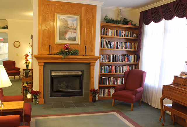 A cozy living room area with a wooden fireplace mantle, a framed landscape painting above it, and two red armchairs. To the right of the fireplace is a tall bookshelf filled with books. A window with sheer white curtains and a maroon valance lets in natural light. A wooden piano is partially visible on the right side, and a table with a lamp and flowers is on the left.