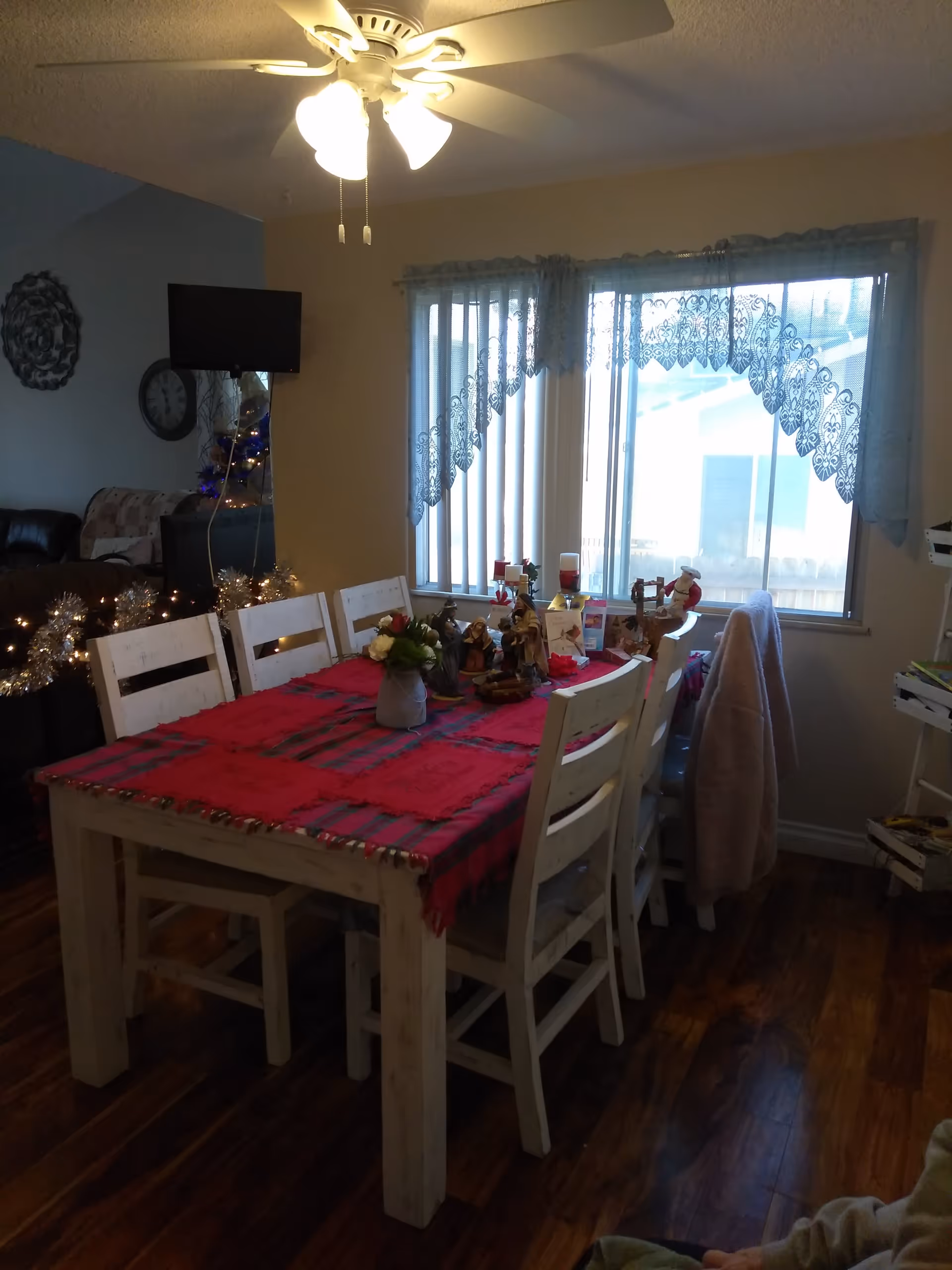 A dining room with a wooden table covered by a red tablecloth and six white wooden chairs. The table is decorated with various figurines, candles, and a small flower arrangement. A large window with lace curtains allows natural light into the room. A ceiling fan with lights is on above the table. In the background, part of a living room with a wall clock, a mounted TV, and a decorated Christmas tree is visible.