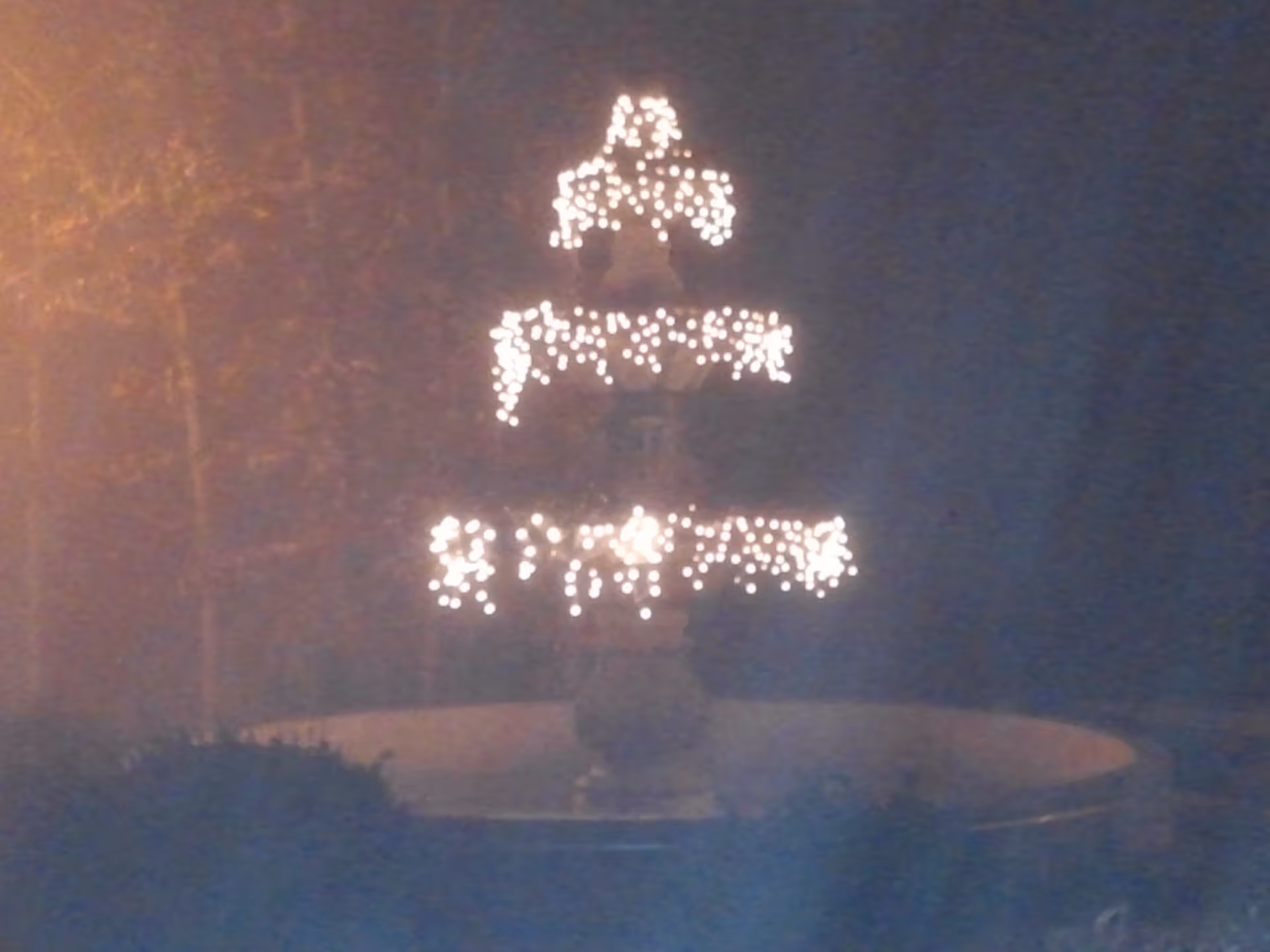 A multi-tiered outdoor fountain decorated with string lights glowing in the dark, surrounded by bushes and trees.