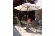 Outdoor patio area with a round glass table, four chairs, and a large white umbrella providing shade. There is a potted plant with red flowers on the table, and trees and part of a building are visible in the background.