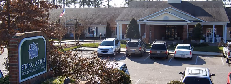 Front exterior view of Spring Arbor of Apex residential assisted living facility with a parking lot in front, several parked cars, and an American flag on a flagpole near the entrance.