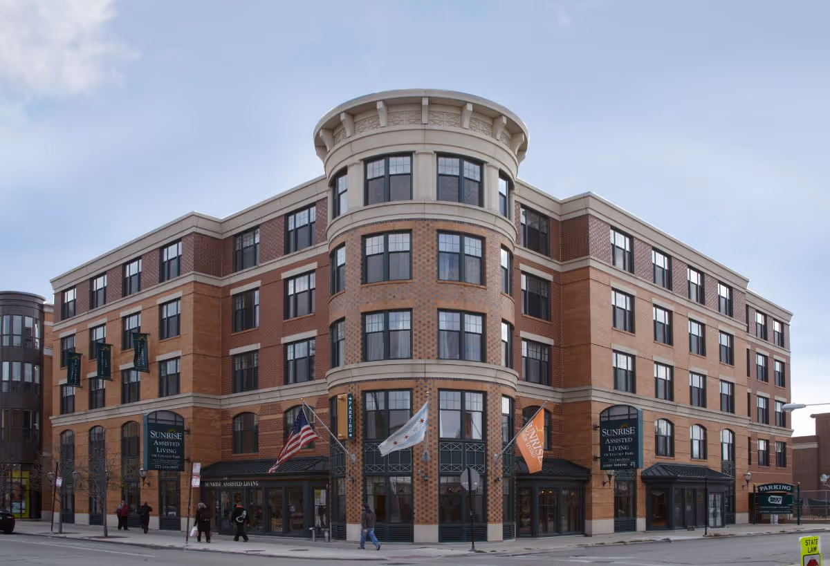Exterior view of a multi-story brick building with large windows and rounded corner section, displaying flags and signs for Sunrise Assisted Living of Lincoln Park. Several people are walking on the sidewalk in front of the building.