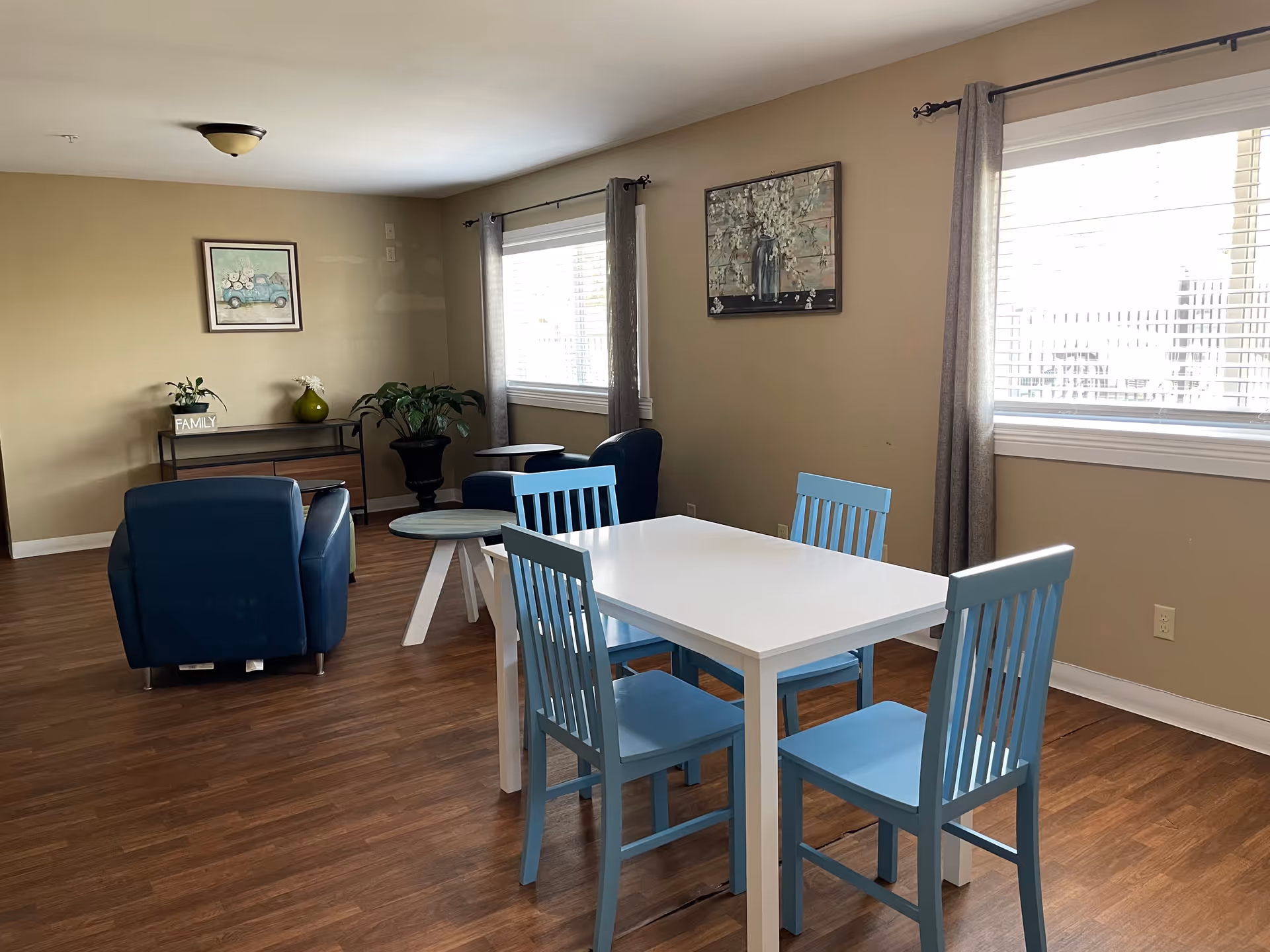 A bright room with wooden flooring featuring a white rectangular table surrounded by four light blue chairs. In the background, there are two blue armchairs with small round tables beside them, a console table with a green vase and a small plant, and two framed paintings on beige walls. Two large windows with gray curtains allow natural light to fill the space.