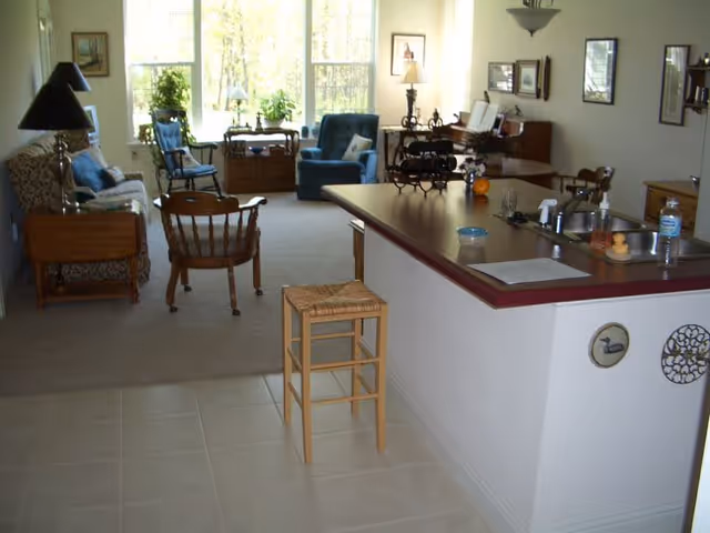 Interior view of a senior living facility showing a combined living room and kitchen area. The living room has several chairs, a couch, a lamp, and large windows letting in natural light. The kitchen area features a counter with a sink, a wooden stool, and various items on the counter including a water bottle and a small bowl.
