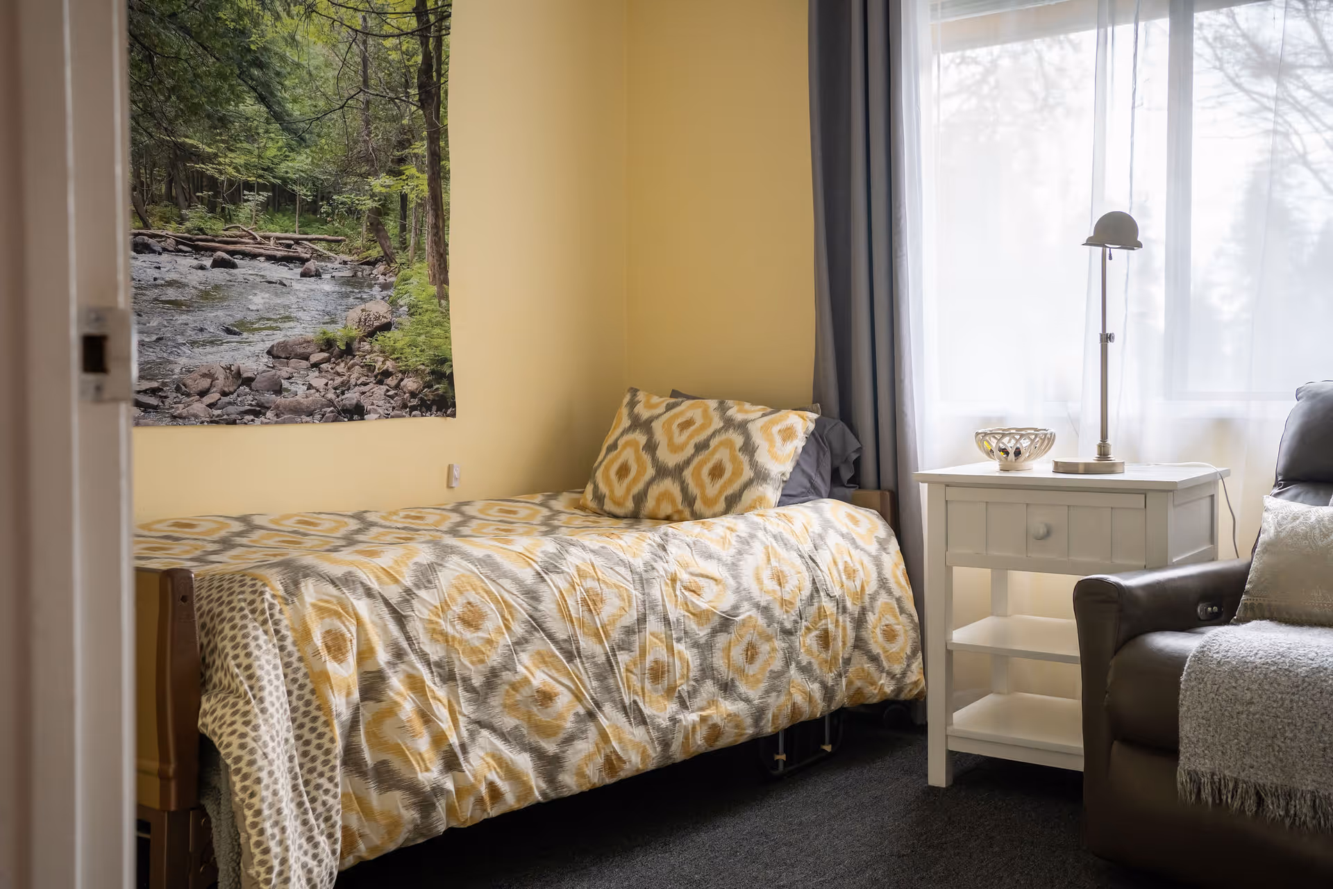 A cozy bedroom with a single bed covered in a yellow and gray patterned comforter and matching pillow. Next to the bed is a white nightstand with a lamp and decorative bowl. A large window with sheer white curtains lets in natural light. A brown armchair with a gray throw blanket is partially visible on the right side. A wall hanging depicting a forest stream scene is on the wall above the bed.