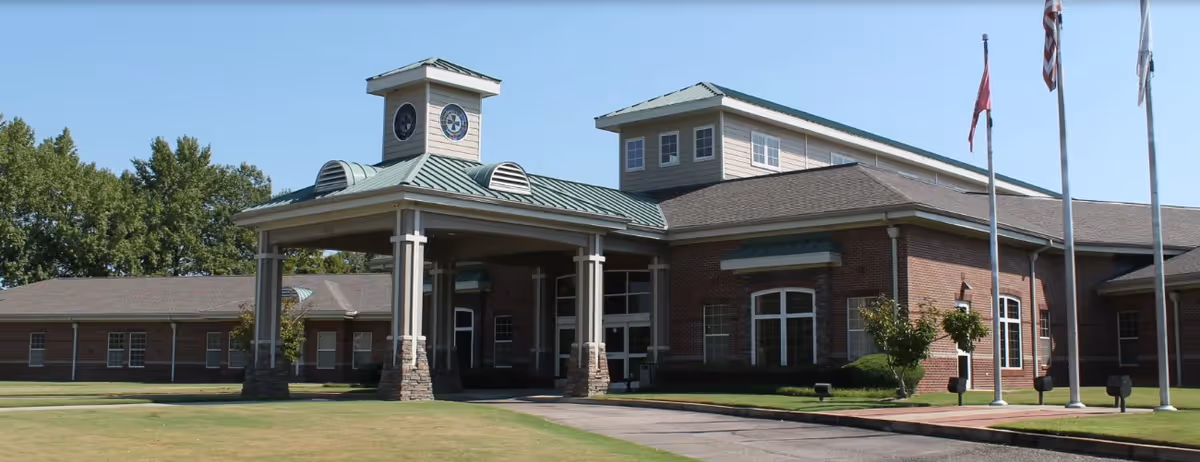 Exterior view of a large brick building with a covered entrance supported by columns and a clock tower above it. The building has multiple windows and a green metal roof. There are three flagpoles with flags in front of the building, and a well-maintained lawn surrounds the area under a clear blue sky.
