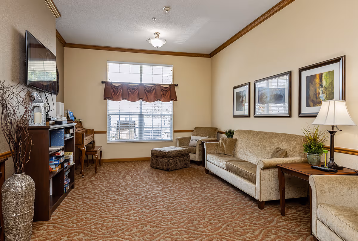 A comfortable living room with a sofa, armchair and ottoman, TV, side tables, and a window with a valance.