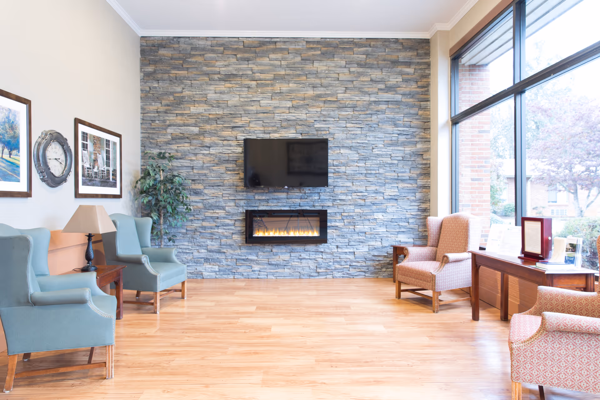 Bright lounge with armchairs, a wall-mounted TV and an electric fireplace against a stone accent wall, hardwood floors and large windows.