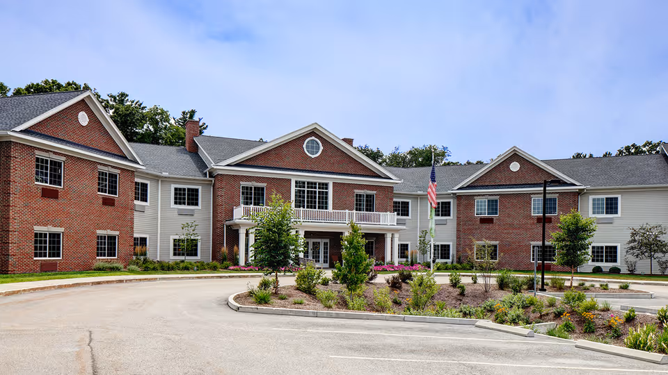 Front exterior view of a two-story senior living facility building with red brick and white siding, a circular driveway, landscaped garden with shrubs and flowers, and an American flag on a flagpole.