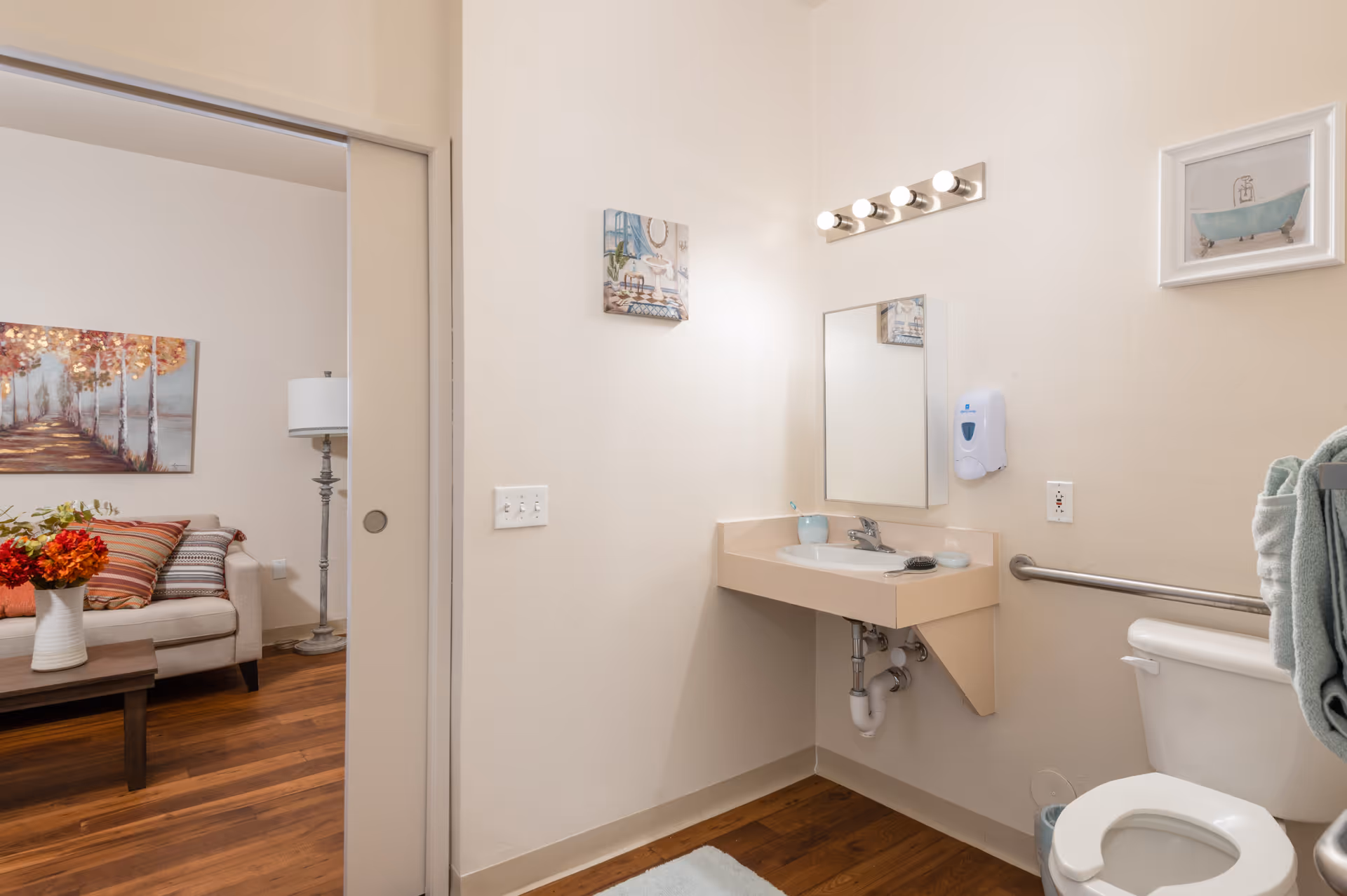 A clean and accessible bathroom with a wall-mounted sink, mirror, and light fixture above. There is a toilet with a grab bar and a towel hanging on the side. The bathroom has light-colored walls and wood flooring. Through an open sliding door, a living room area with a couch, floor lamp, and wall art is visible.