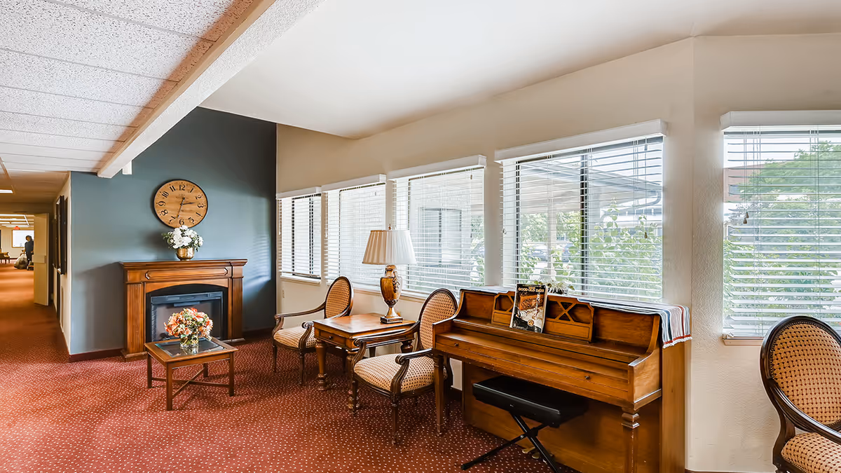 Communal sitting area with a piano, upholstered chairs, a small table, and a decorative fireplace beside large windows.