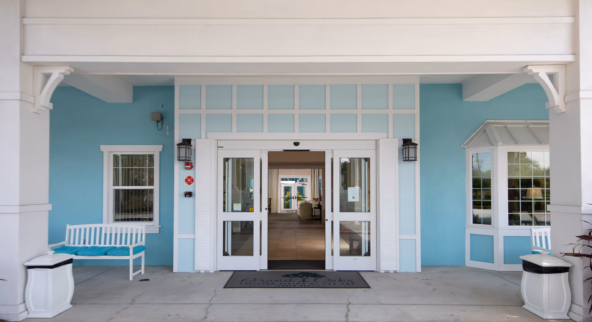 Entrance of a building with light blue walls and white trim, featuring double glass doors. There are white benches with blue cushions on either side of the entrance and two white trash bins. The floor mat in front of the doors reads 'Grand Oaks of Jensen Beach'.