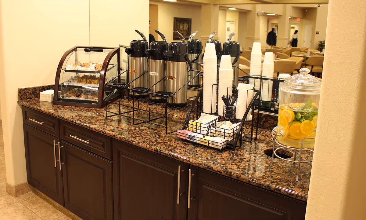 A coffee and refreshment station with multiple stainless steel coffee dispensers, stacks of white disposable cups, a container with lemon and mint infused water, and a display case with assorted pastries on a granite countertop in a senior living facility.