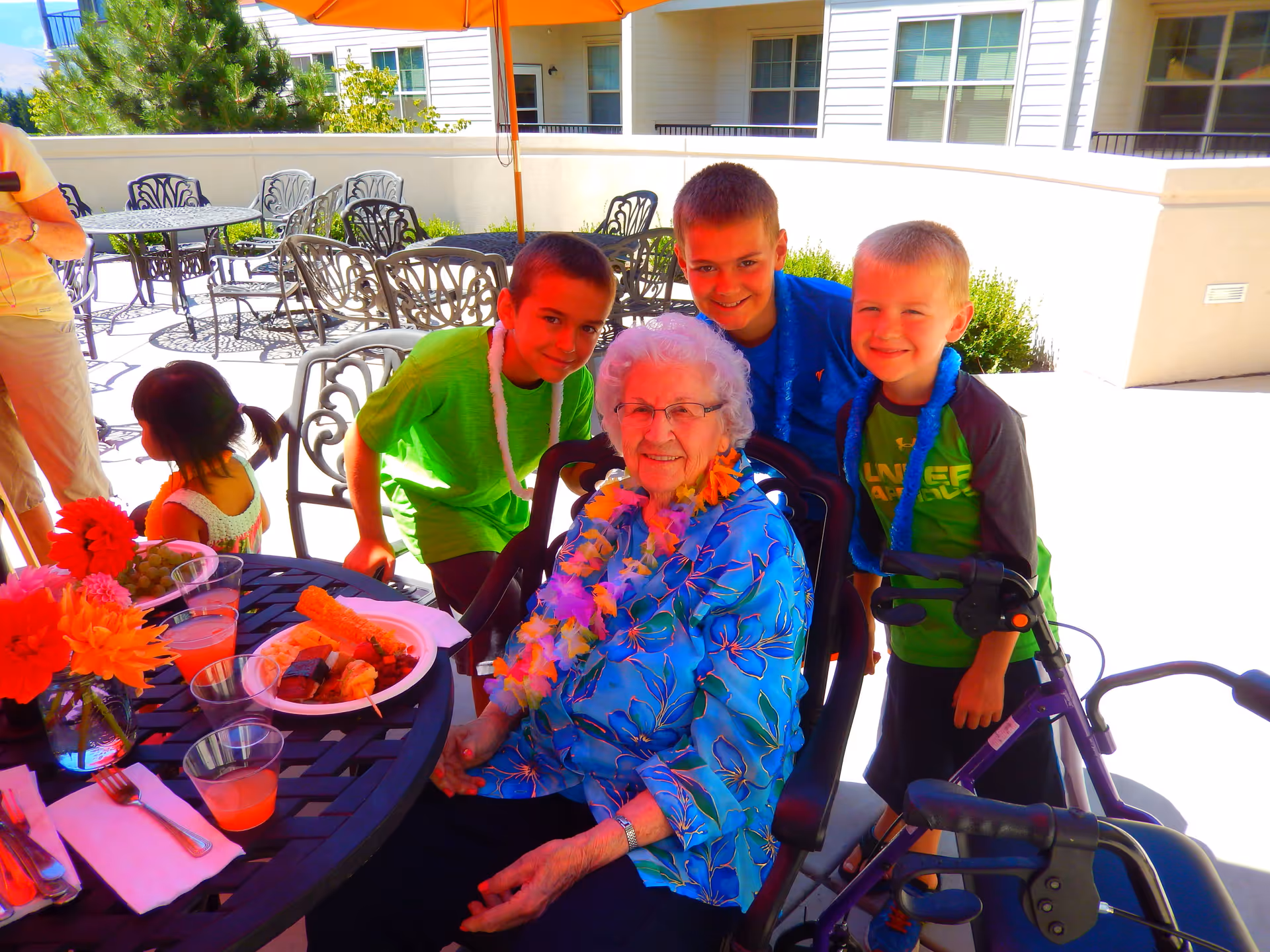An elderly woman wearing glasses and a blue floral shirt sits in a wheelchair at an outdoor patio table decorated with flowers and food. She is surrounded by three smiling young boys, two of whom are wearing leis. Another child and an adult are partially visible in the background. The setting is a sunny outdoor patio with tables, chairs, and an orange umbrella.