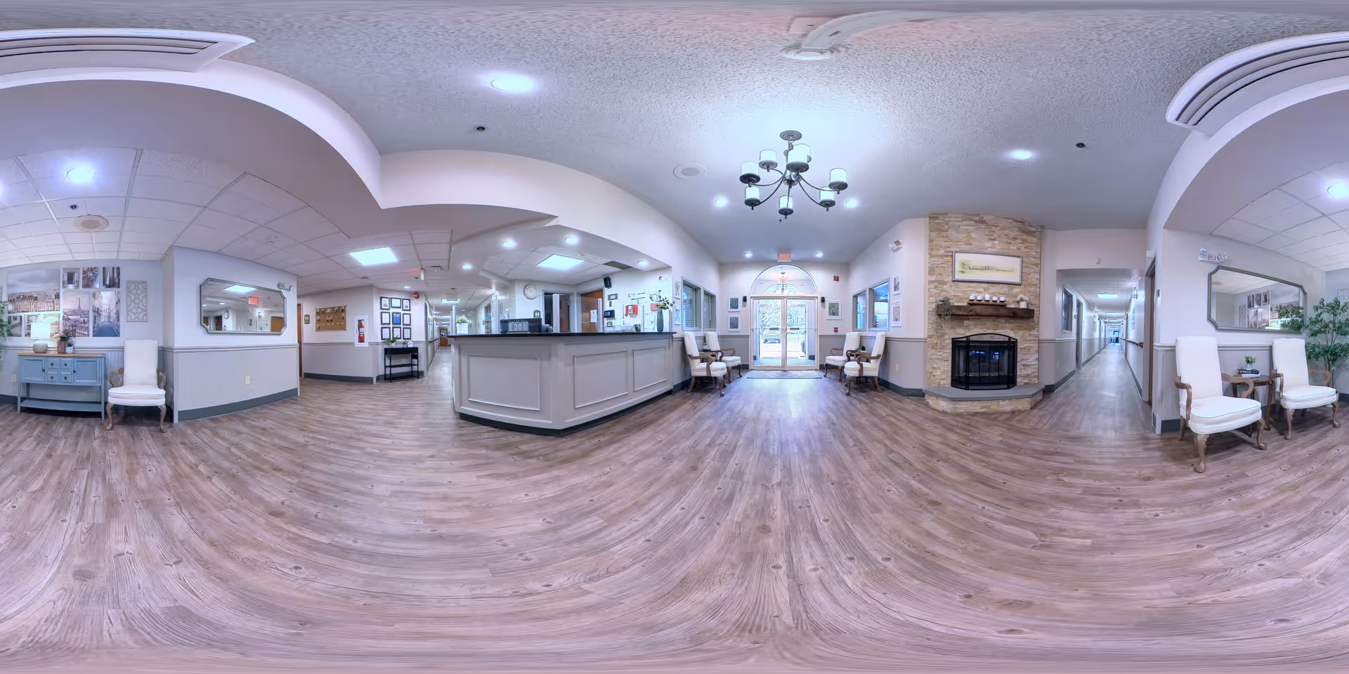 Wide panoramic view of a senior living facility's interior lobby area with a reception desk in the center, seating areas with chairs on both sides, a stone fireplace on the right wall, framed artwork, and a chandelier hanging from the ceiling. The floor is wood-style, and there are hallways leading to other parts of the facility.