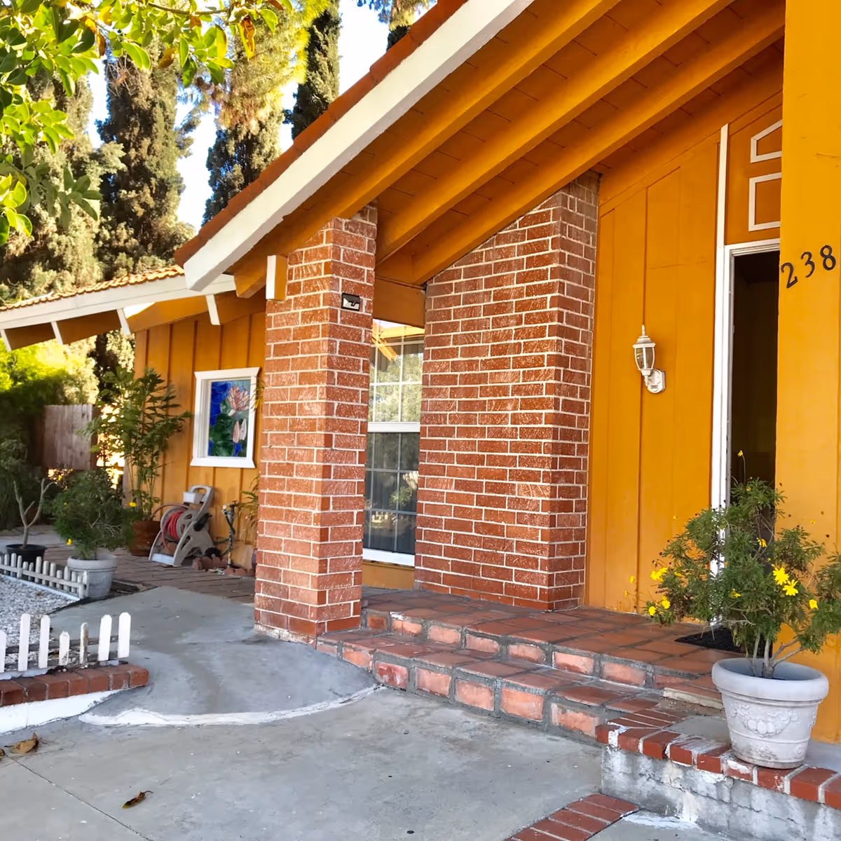 Front exterior view of a yellow building with brick pillars and steps leading to an open door. There are potted plants and trees around the entrance, and a small white picket fence borders a garden area.