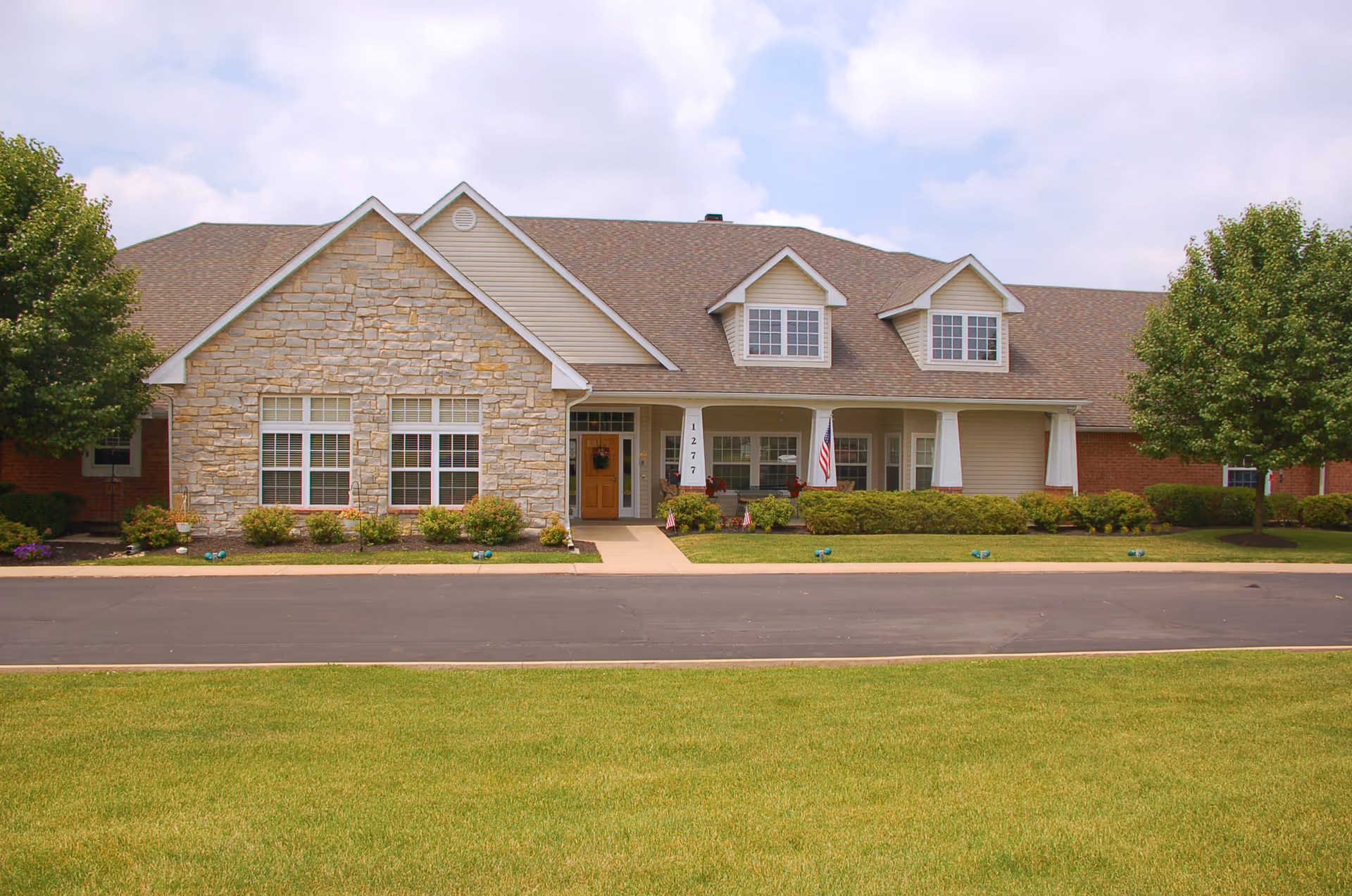 Front view of a single-story stone-and-siding senior living building with a covered porch, manicured lawn, and an American flag.
