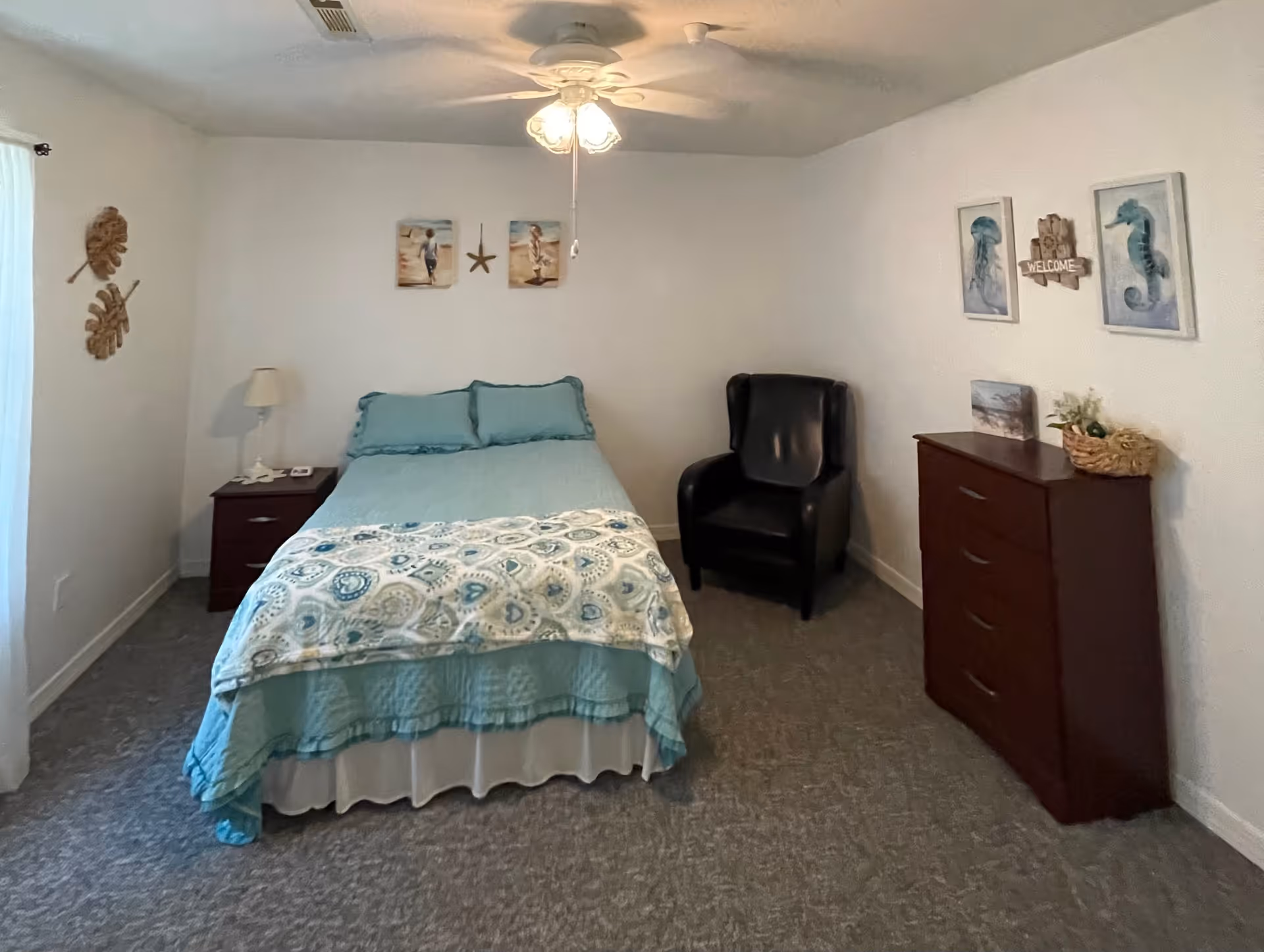 A bedroom with a bed covered in blue and white bedding, a nightstand with a lamp on the left, a black armchair in the corner, and a wooden dresser on the right. The walls are decorated with beach-themed artwork and a welcome sign.