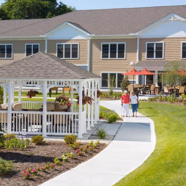 Two elderly women walking on a paved pathway in a well-maintained garden area with a white gazebo adorned with hanging flower baskets. In the background, there is a two-story building with beige siding and multiple windows, along with an outdoor seating area featuring tables with red umbrellas.