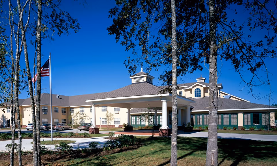 Exterior view of Garden View Assisted Living facility showing a large building with a covered entrance, several windows, and a flagpole with an American flag. Trees and landscaped greenery are visible in the foreground under a clear blue sky.