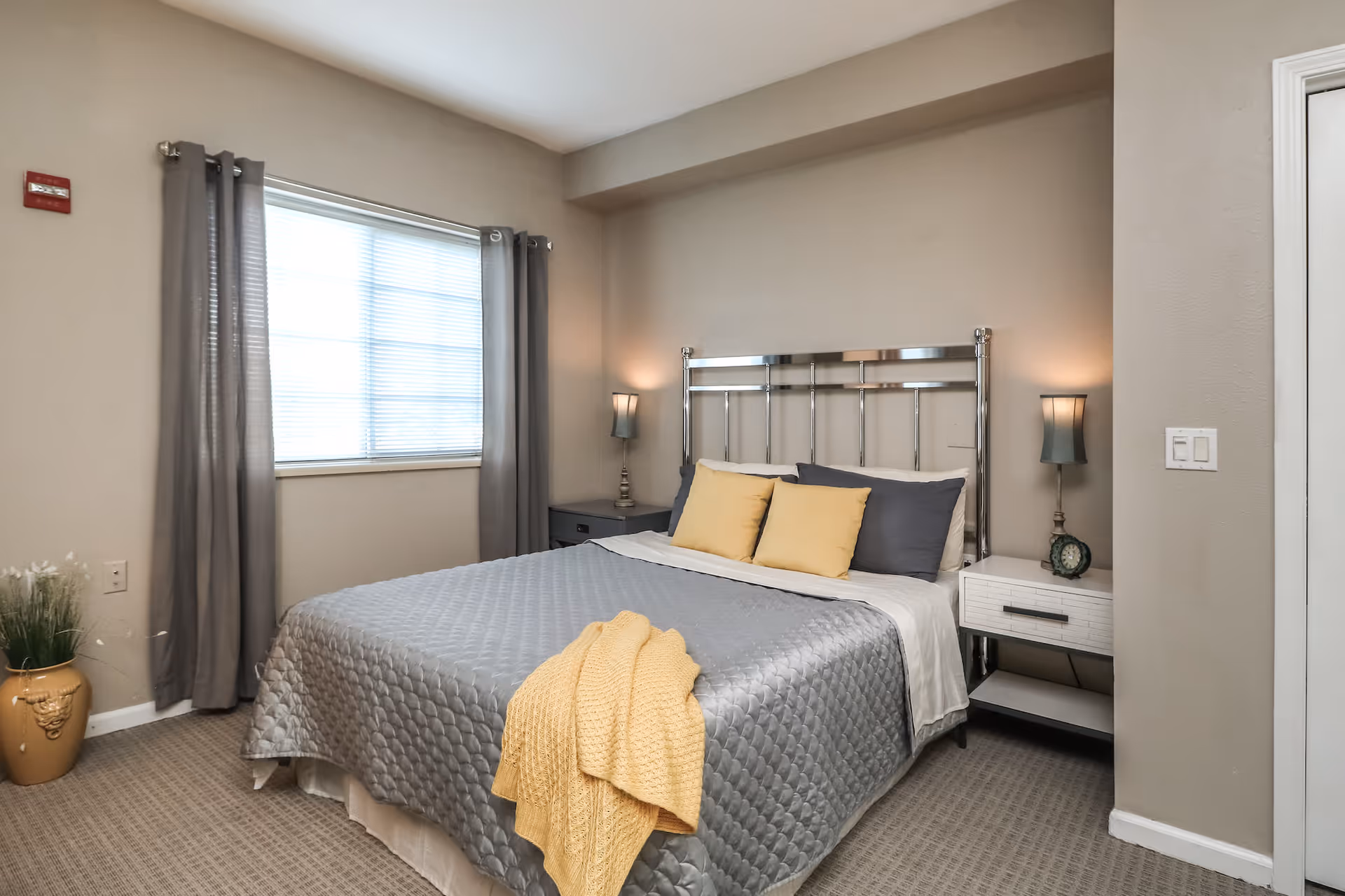 A neatly made bed with a gray quilted bedspread and yellow pillows and throw blanket in a bedroom. The room has beige walls, a window with gray curtains, two bedside tables with lamps, and a small decorative plant in a vase on the floor.