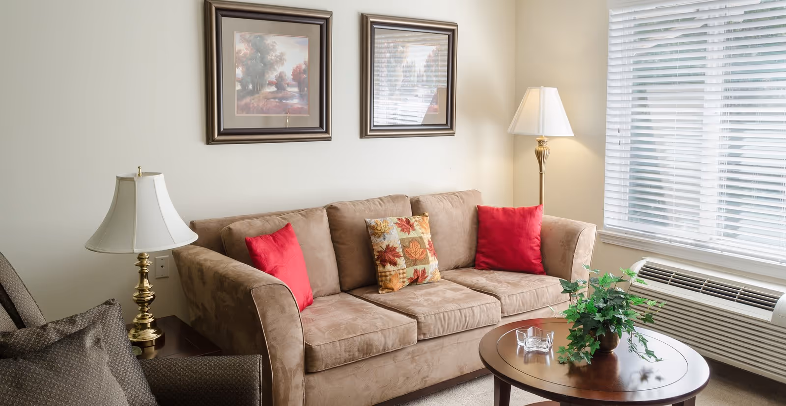 A cozy living room with a beige sofa adorned with two red pillows and one decorative pillow with autumn leaves. There are two framed landscape paintings on the wall above the sofa. A round wooden coffee table with a green potted plant and a glass ashtray sits in front of the sofa. To the left is a side table with a brass lamp and a cushioned armchair. A tall floor lamp stands in the corner near a window with white blinds, and an air conditioning unit is below the window.