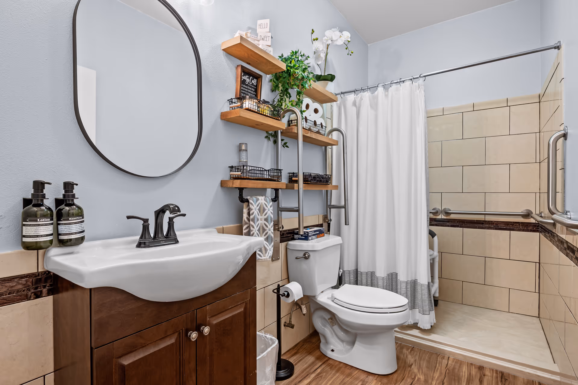 Bright bathroom with a sink and mirror, wooden vanity, shelving over the toilet, and a tiled walk-in shower with grab bars.