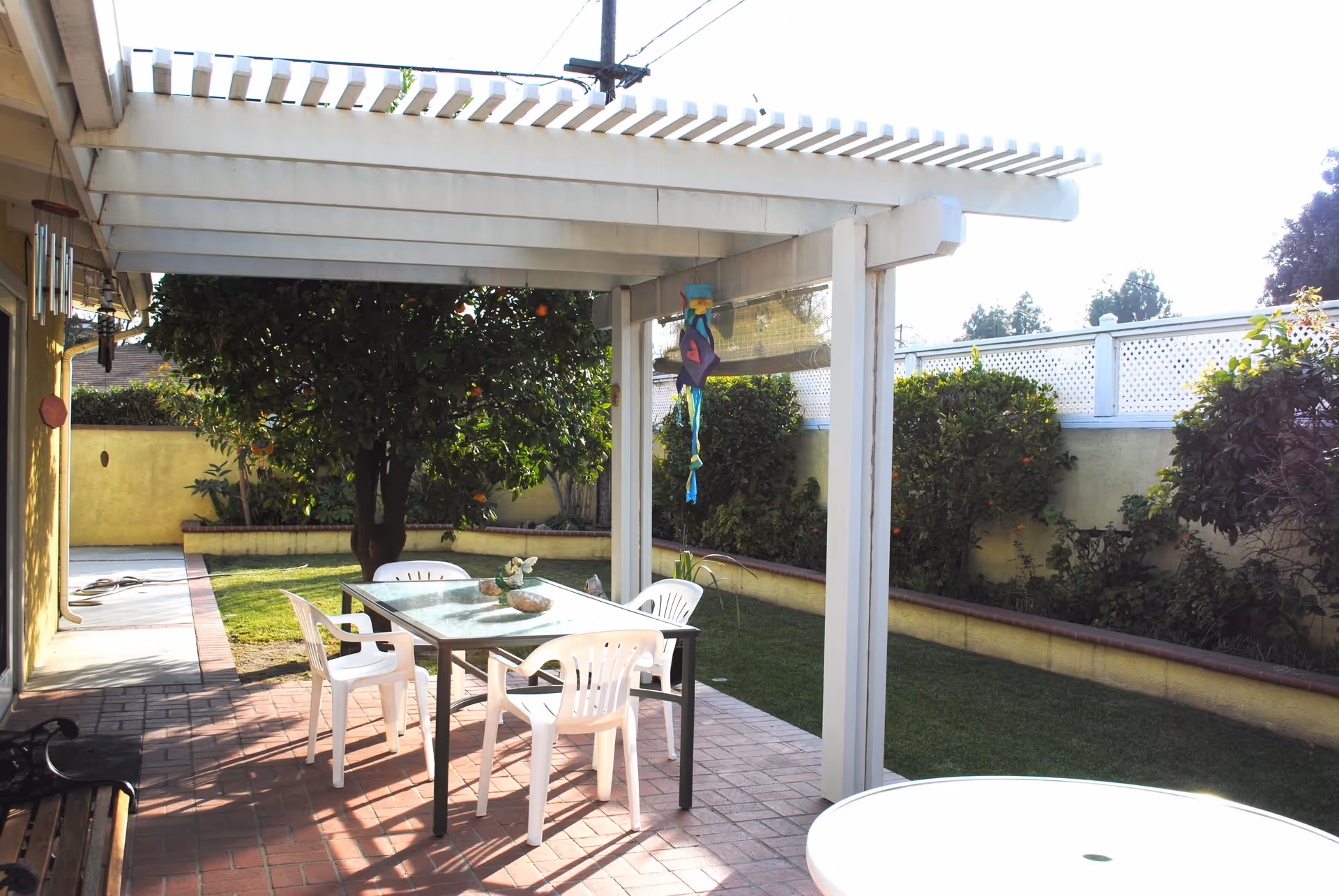 Outdoor patio area with a white pergola overhead, a glass-top table surrounded by four white plastic chairs, a green lawn, and various bushes and trees along a yellow wall with white lattice fencing on top.