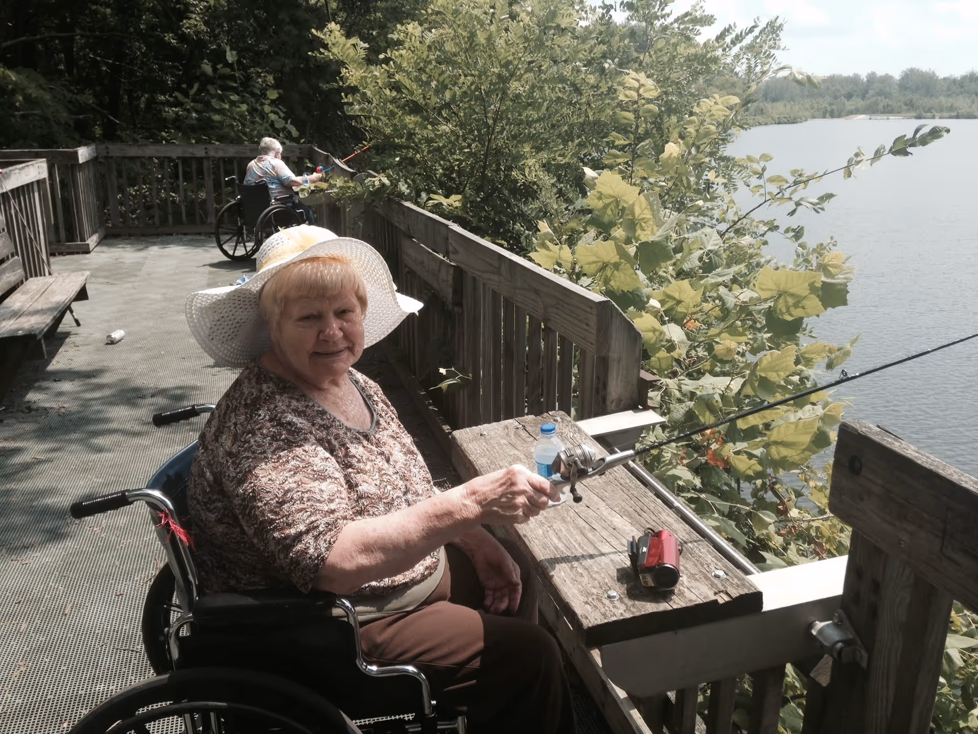 An elderly woman in a wheelchair wearing a white sun hat and patterned shirt is fishing from a wooden dock overlooking a lake. Another elderly person in a wheelchair is fishing further down the dock. The dock is surrounded by green foliage and trees.