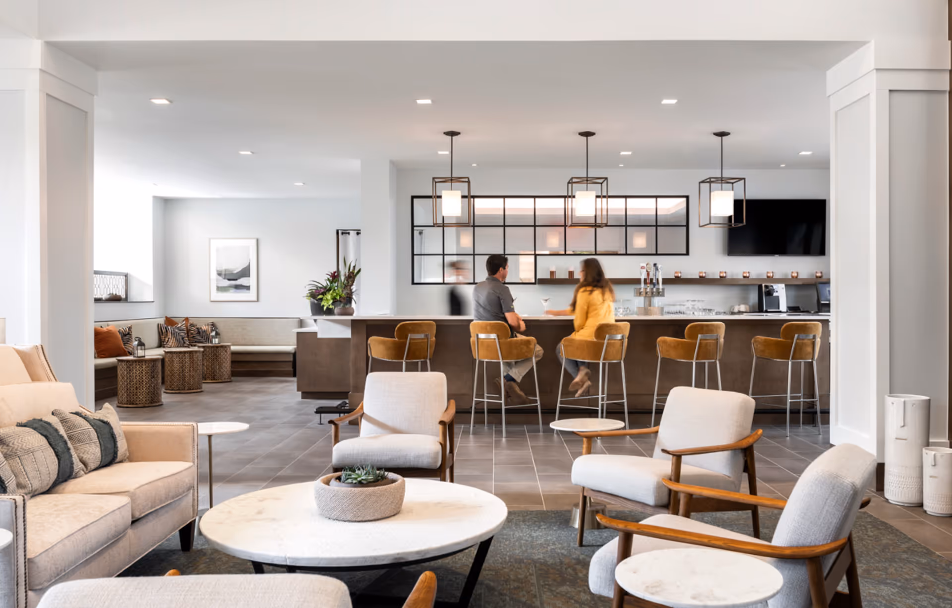 A modern senior living facility common area with a seating arrangement including a beige sofa, armchairs, and round marble tables in the foreground. In the background, there is a bar with four brown bar stools where two people are seated and conversing. The space is well-lit with pendant lights and has a clean, contemporary design with neutral tones.
