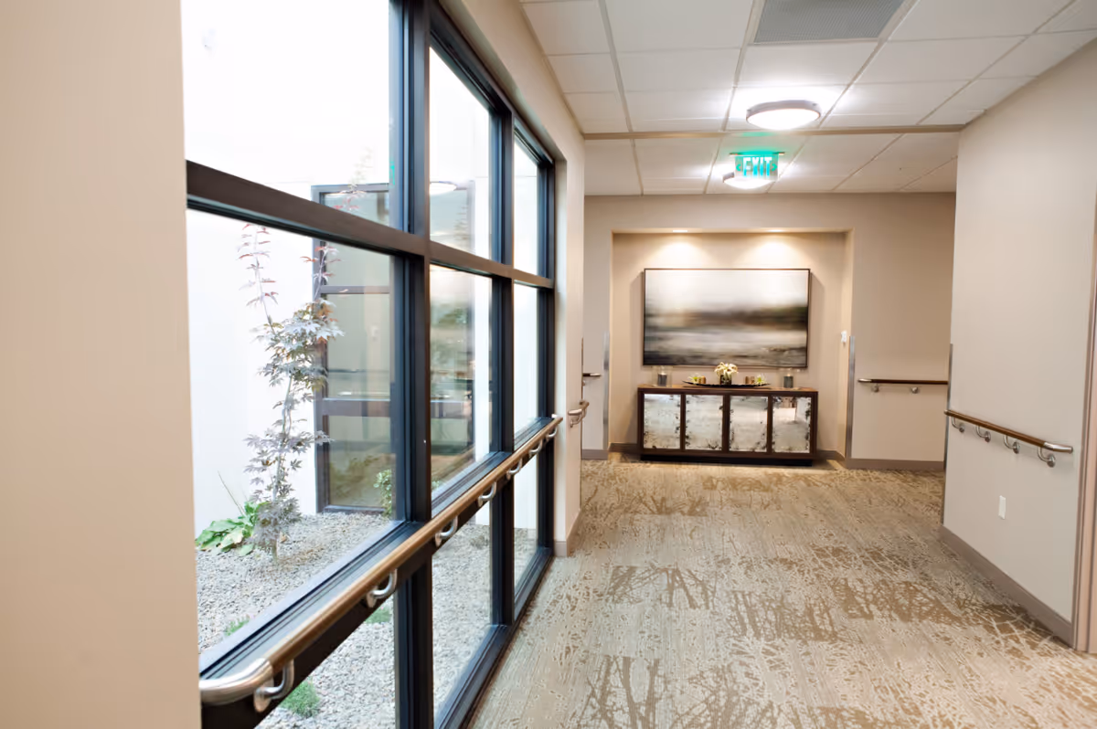 A well-lit hallway in a senior living facility with large windows on the left side showing an outdoor garden area with plants. The hallway has beige walls, handrails on both sides, and a patterned carpet floor. At the end of the hallway, there is a decorative cabinet with a large framed artwork above it and small decorative items on top.