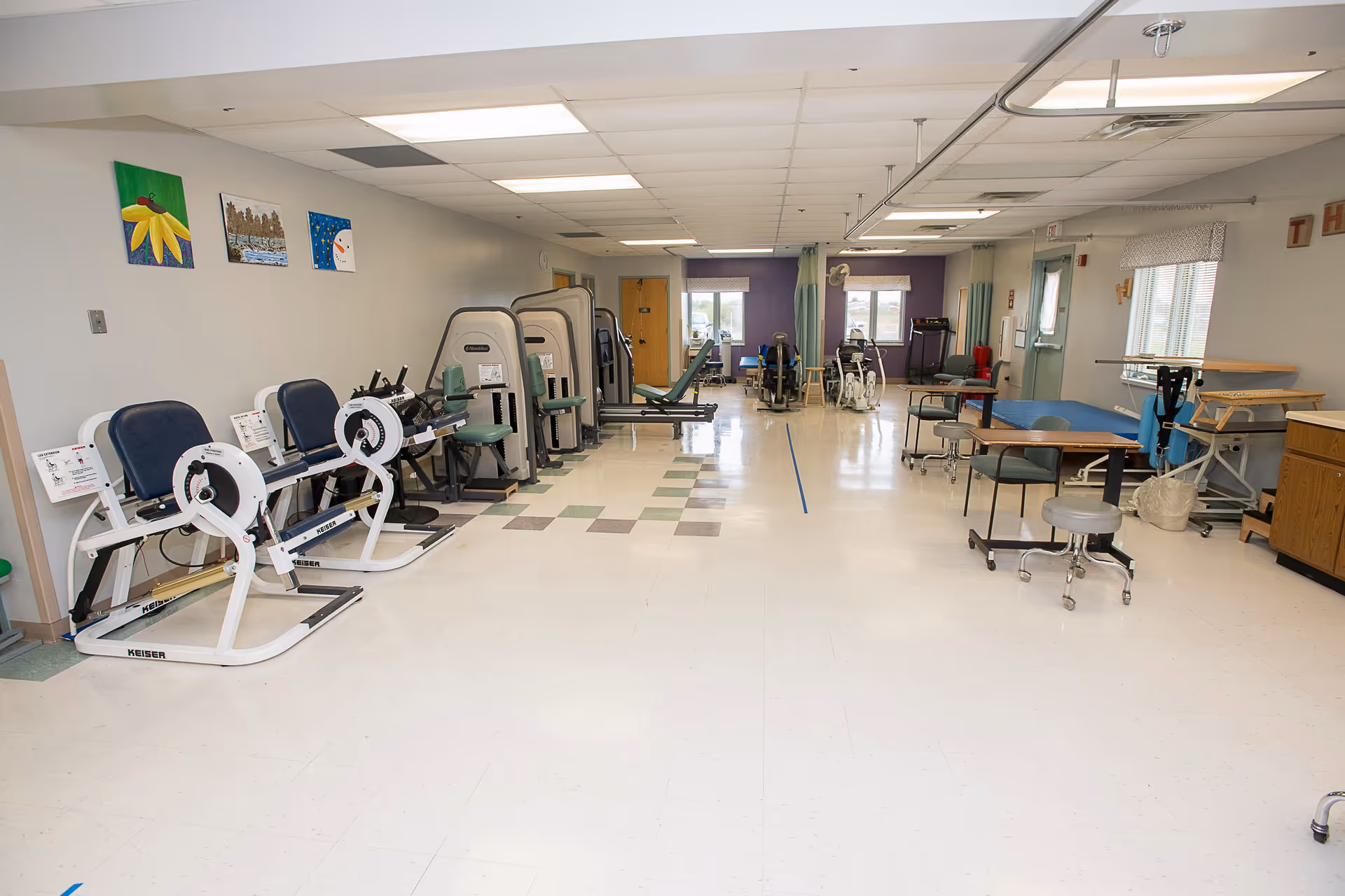 A spacious rehabilitation or physical therapy room with exercise equipment including leg press machines, stationary bikes, and therapy tables. The room has a tiled floor with some colored tiles, white walls with a few paintings, and windows letting in natural light.
