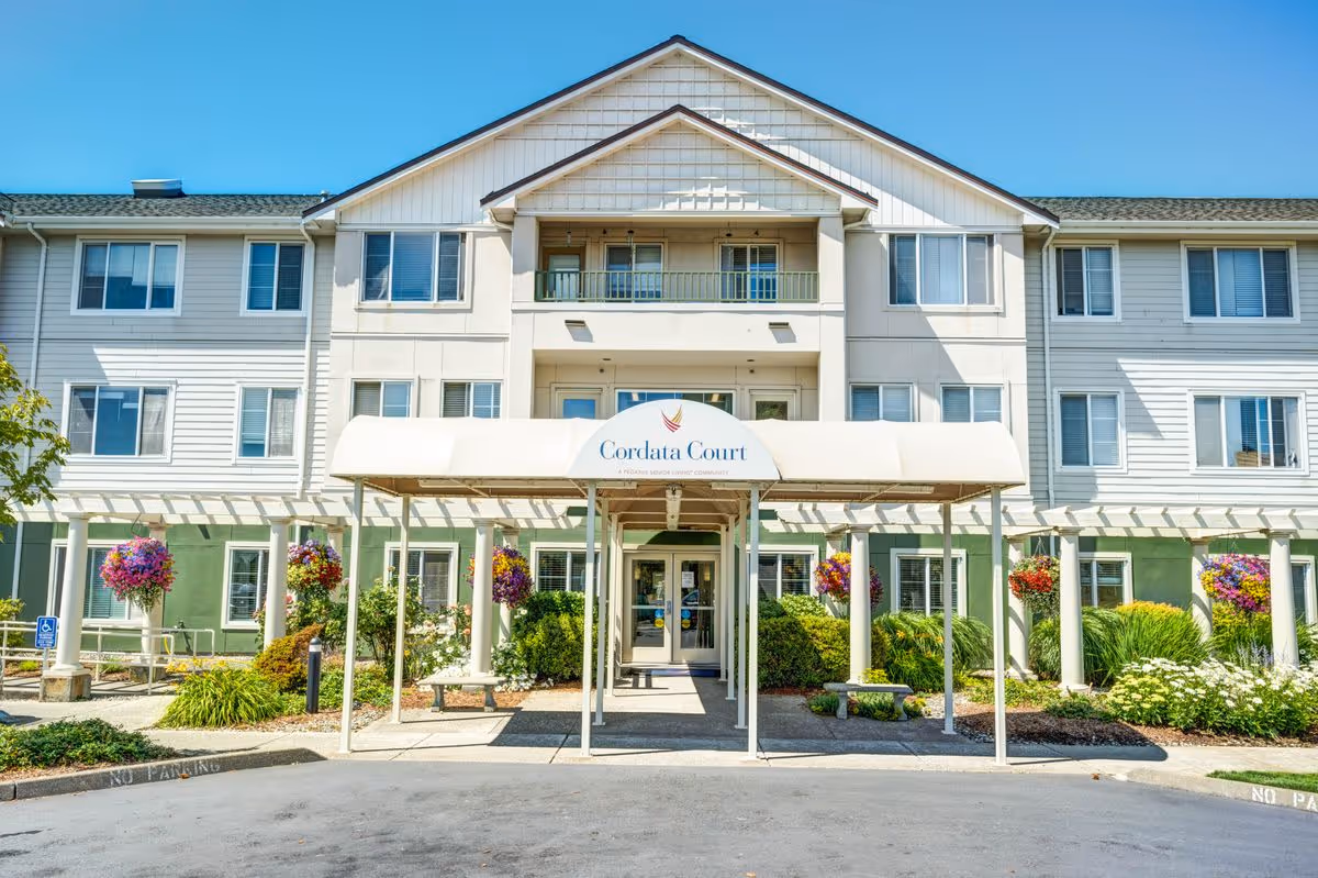 Front exterior view of Cordata Court, a multi-story senior living facility with a covered entrance, hanging flower baskets, and well-maintained landscaping under a clear blue sky.