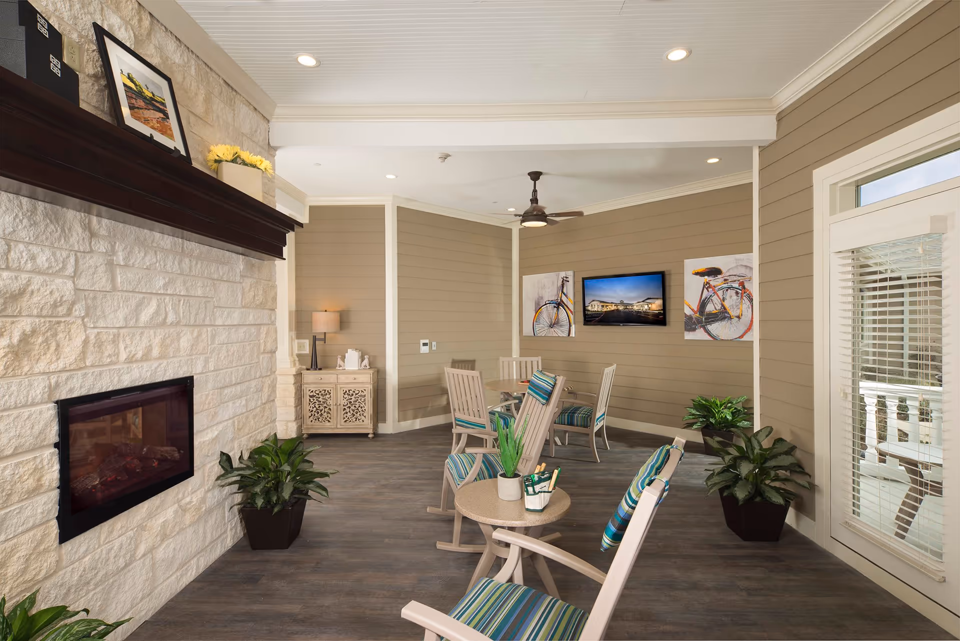 A cozy indoor common area with beige paneled walls and a white stone fireplace. The room features several cushioned rocking chairs with striped blue and green cushions arranged around small tables. There are potted plants placed near the walls, a ceiling fan, a flat-screen TV mounted on the wall, and two bicycle-themed artworks flanking the TV. A small cabinet with a lamp and decorative items is positioned in the corner, and a glass door with blinds leads to an outdoor space.