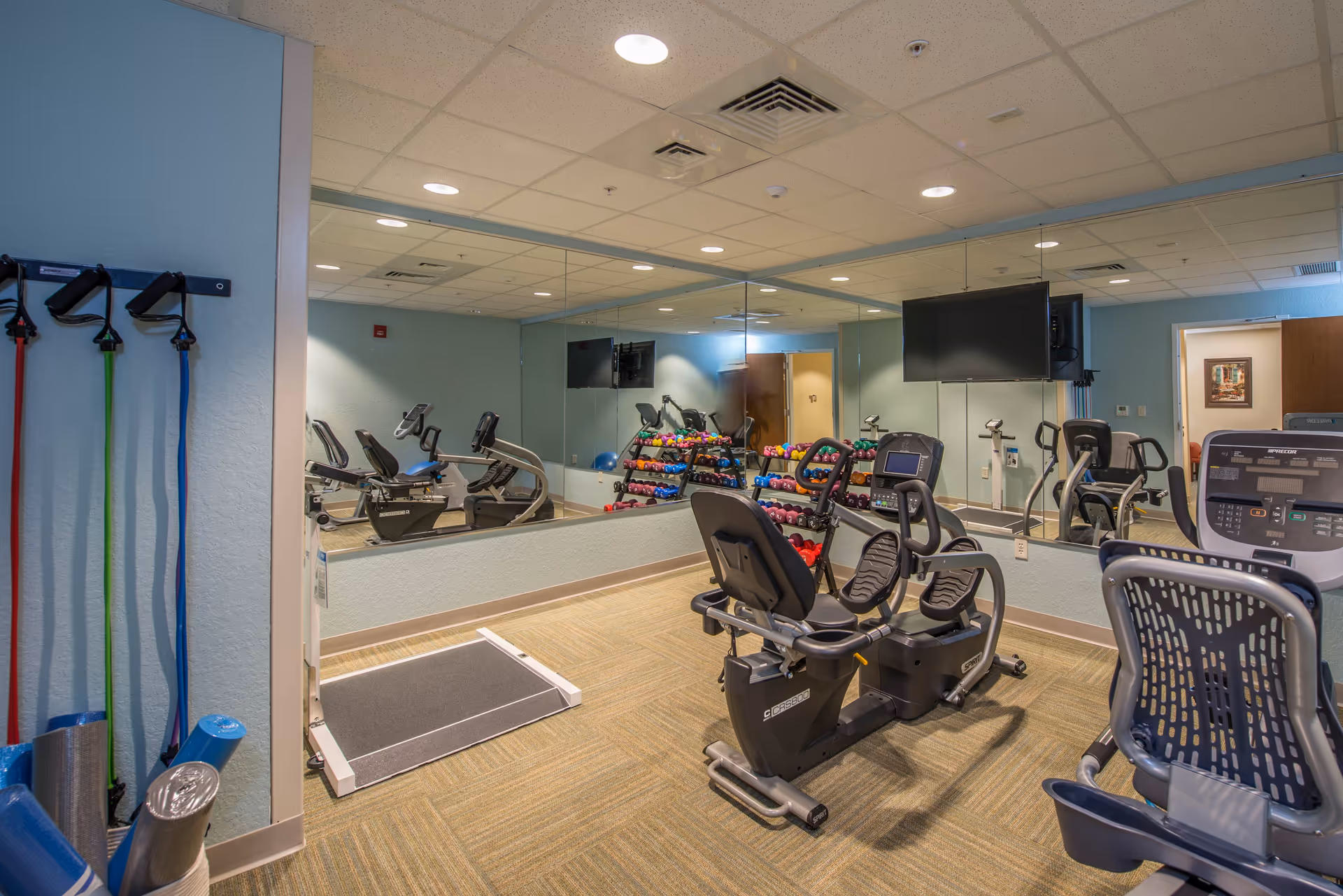A small fitness room with exercise equipment including recumbent bikes, a treadmill, and a rack of colorful dumbbells. The walls are light blue with a large mirror covering one side, reflecting the equipment. Resistance bands and yoga mats are stored on the left wall.
