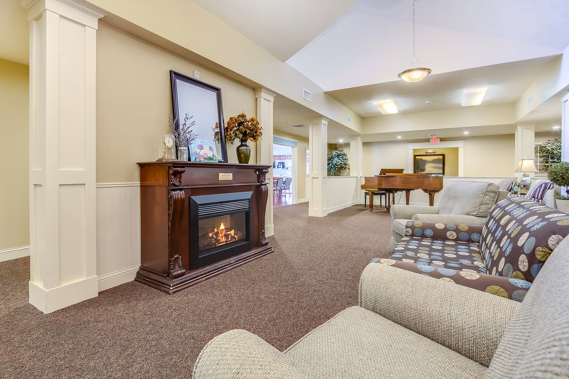 A cozy assisted living facility common area featuring a lit fireplace with a wooden mantel decorated with flowers, a clock, and a framed picture. The room has beige walls, carpeted floors, comfortable upholstered chairs and sofas, and a grand piano in the background. There are plants and soft lighting creating a warm and inviting atmosphere.