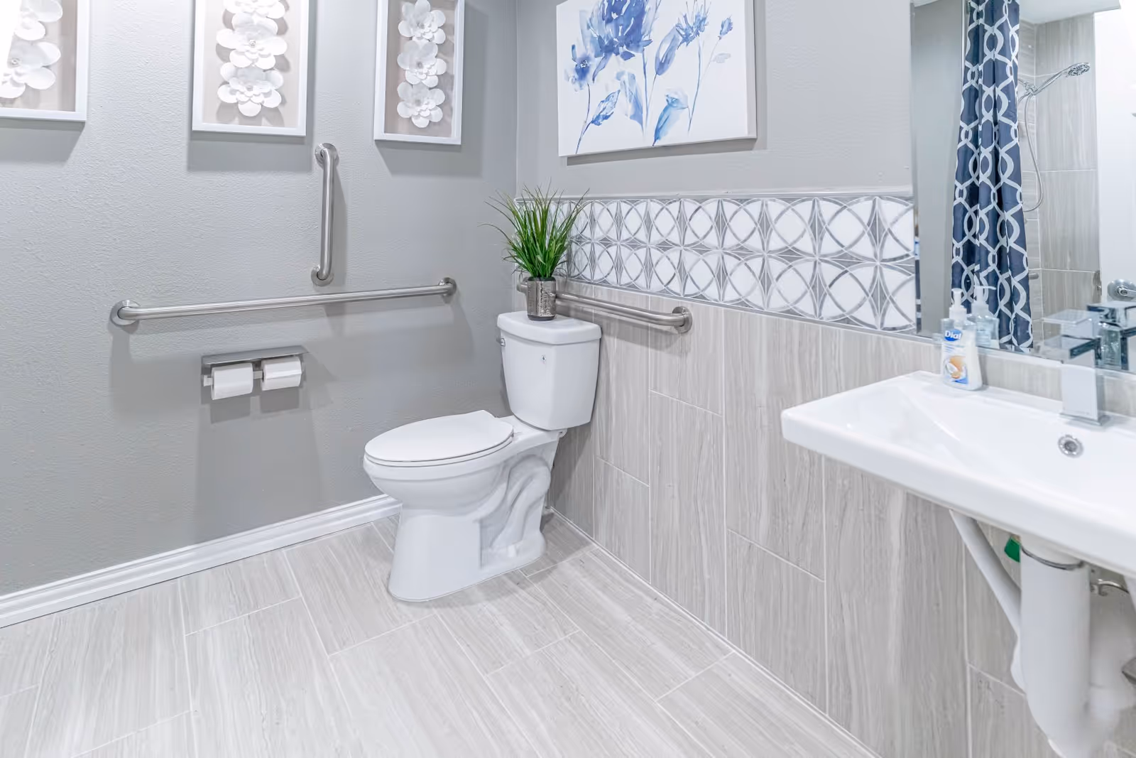 A clean and modern bathroom with a white toilet, grab bars on the walls for accessibility, a white sink with a soap dispenser, decorative wall art, and a shower curtain with a geometric pattern.